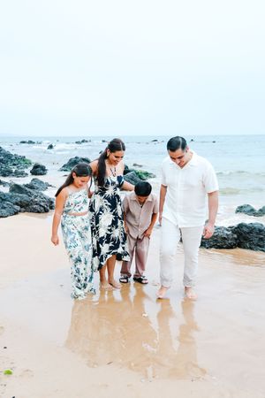 Three people in beachwear stroll along the sandy shoreline near volcanic rocks in Hawaii.