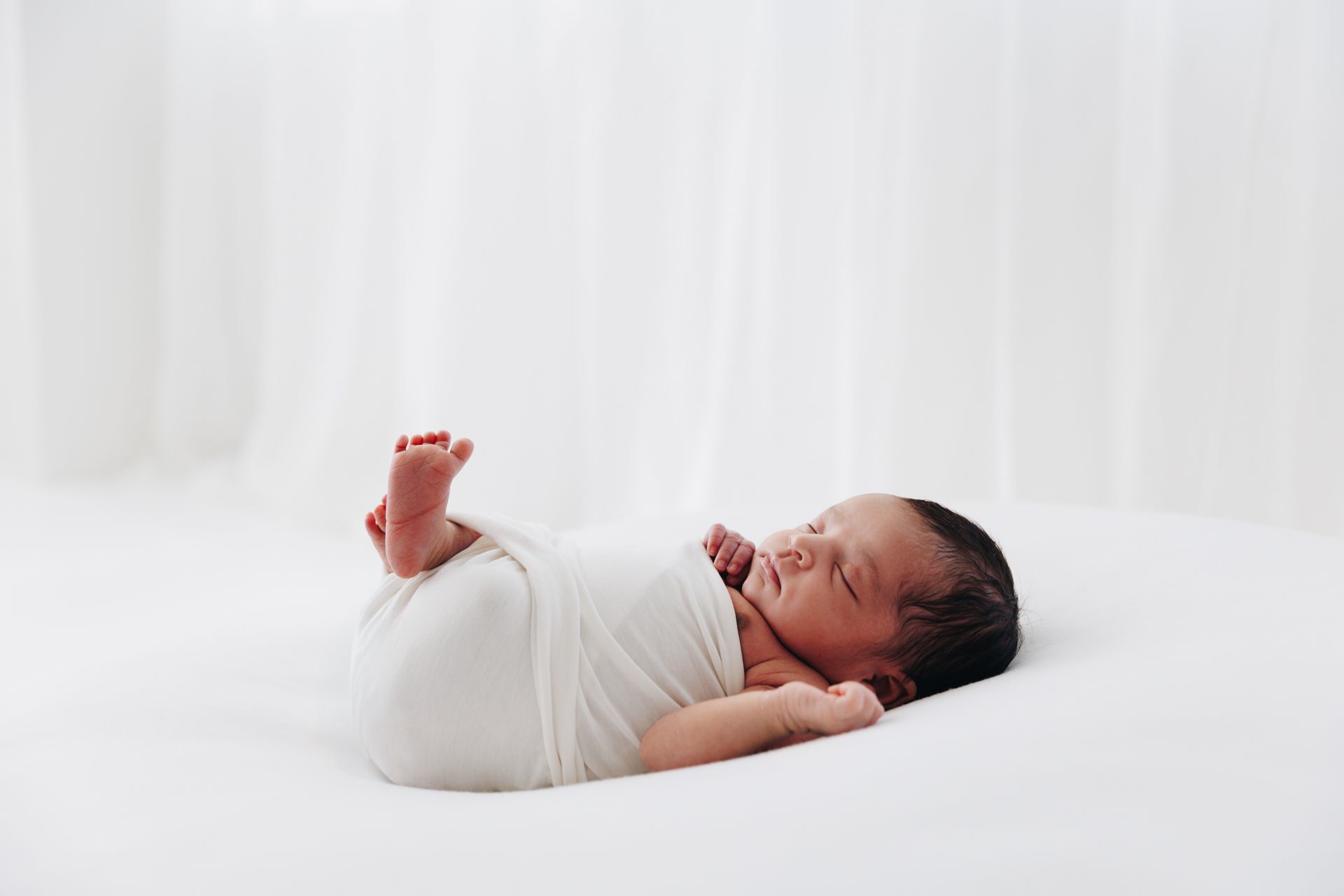 Newborn baby swaddled in white wrap sleeping peacefully on a white surface with soft white background.