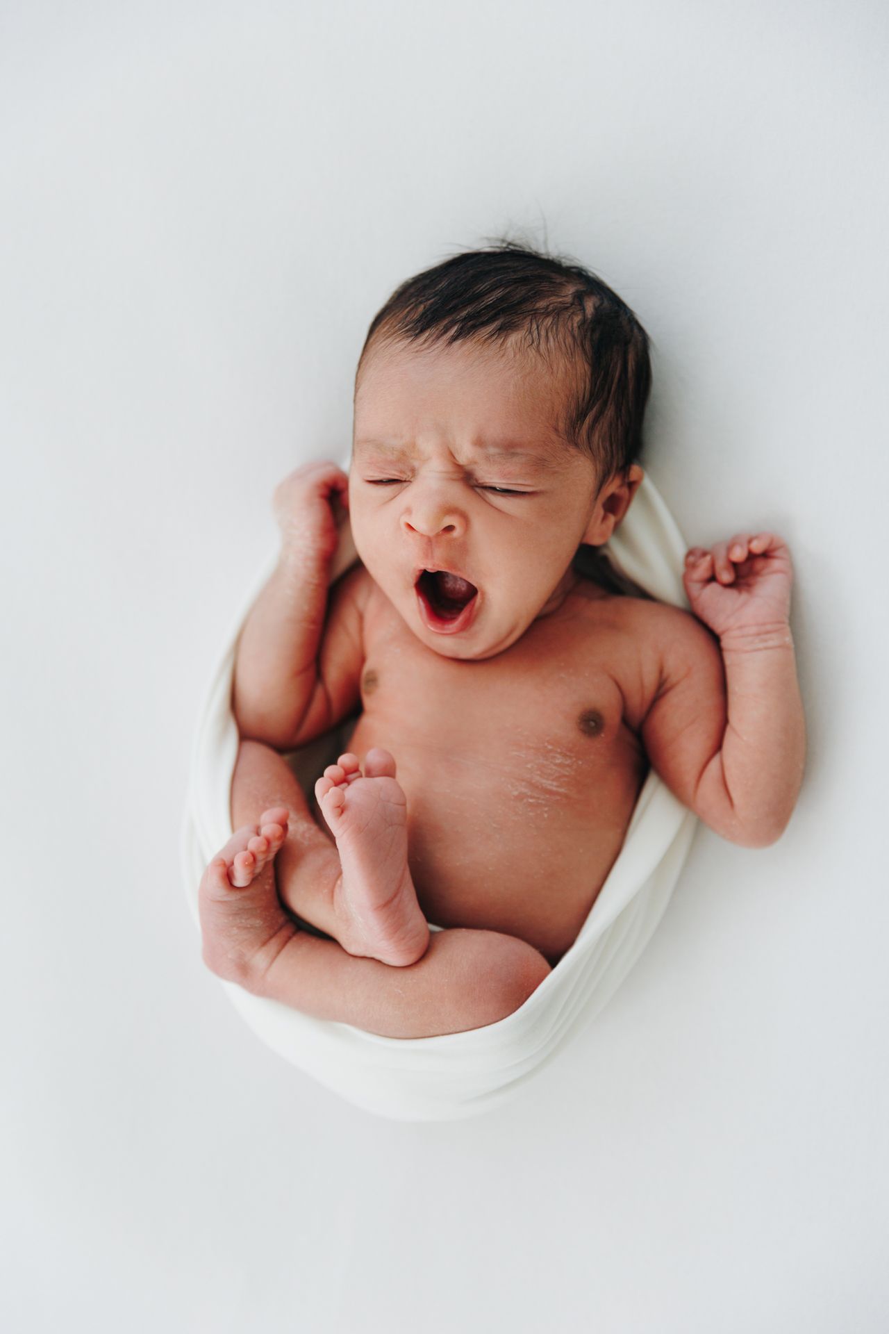 Newborn baby yawning while wrapped in white cloth, lying on white background with arms and legs stretched out.