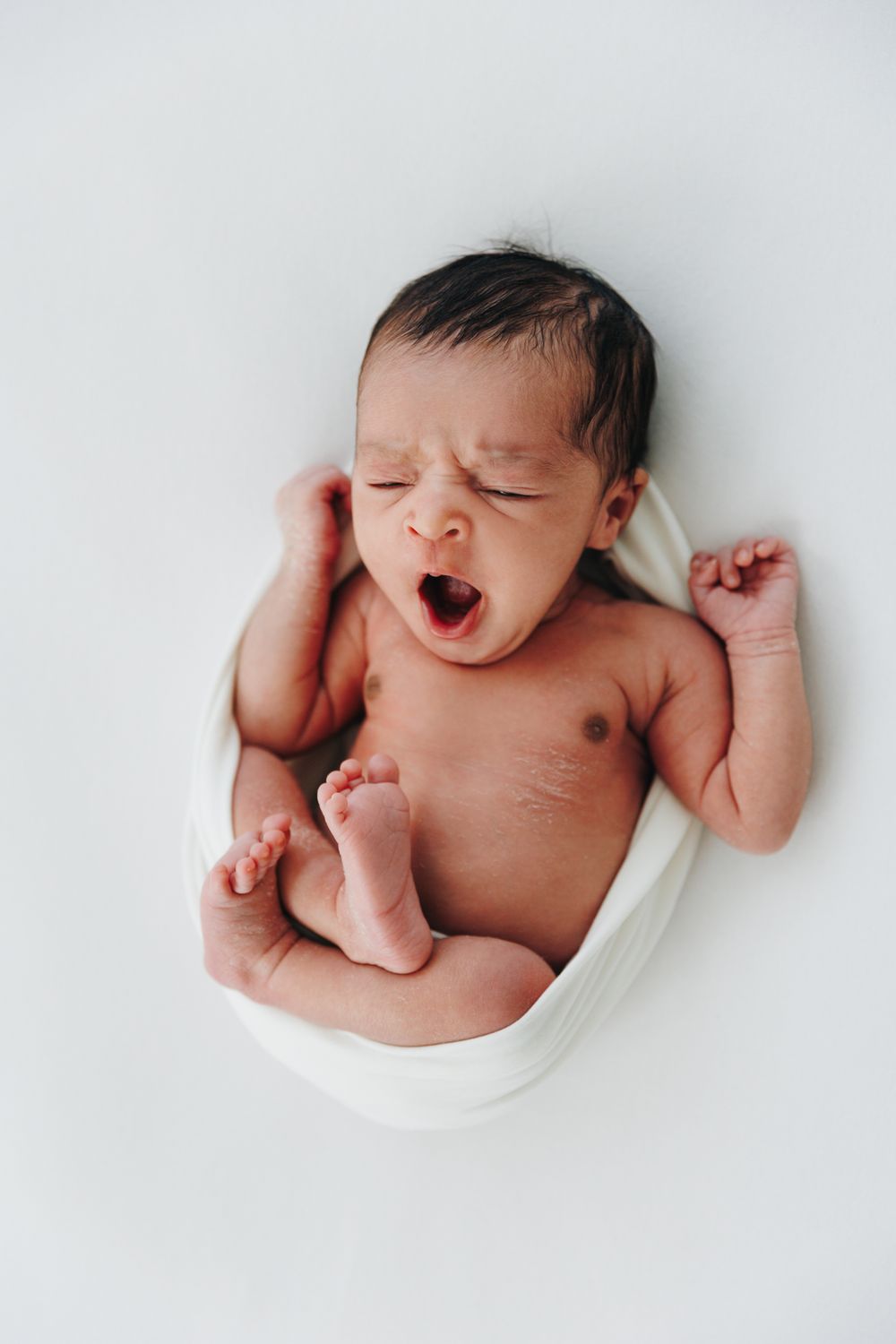 Newborn baby yawning while wrapped in white cloth, lying on white background with arms and legs stretched out.