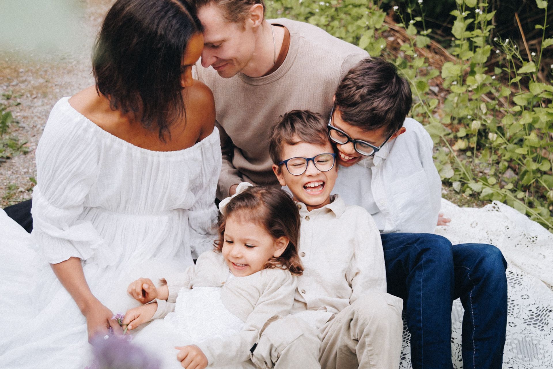 Happy family of five smiling outdoors, parents with three young children sitting together in a bright garden setting.