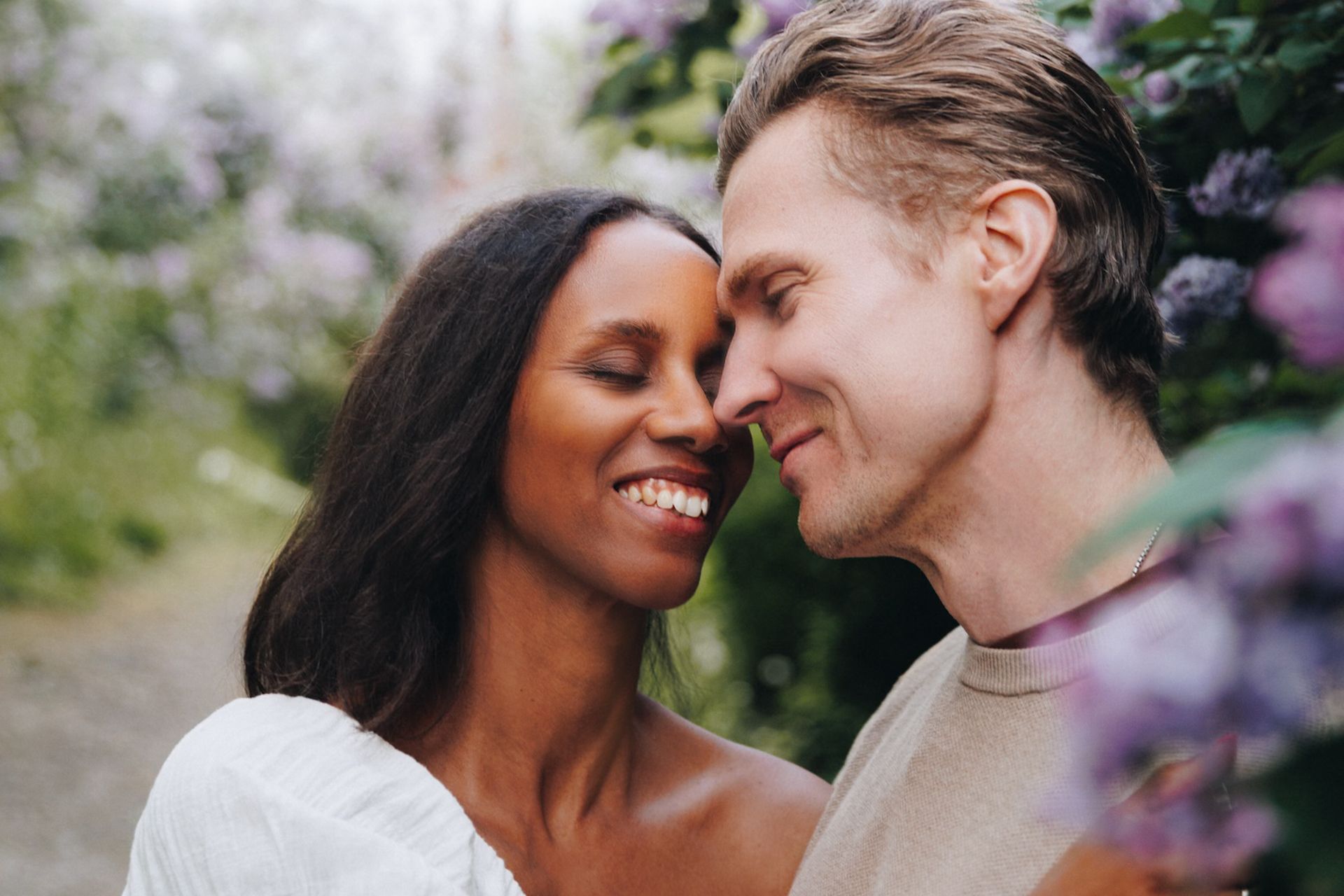 Happy interracial couple smiling and touching foreheads surrounded by blooming purple flowers outdoors.