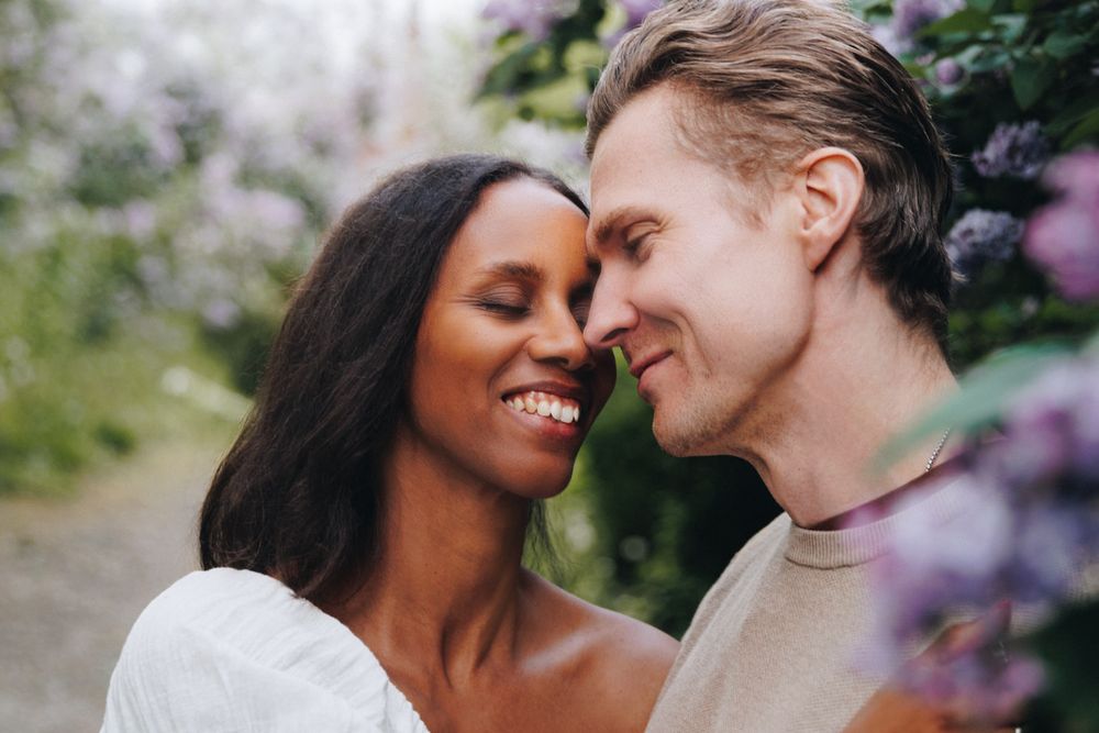 Happy interracial couple smiling and touching foreheads surrounded by blooming purple flowers outdoors.