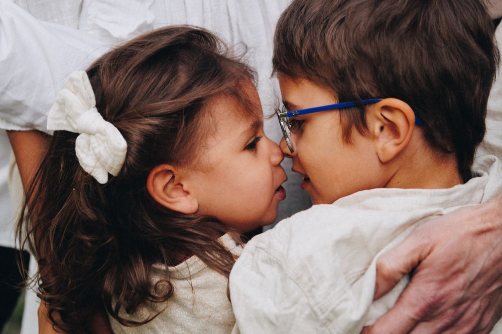 Two young children touching noses affectionately, a girl with a white bow and a boy wearing blue glasses.