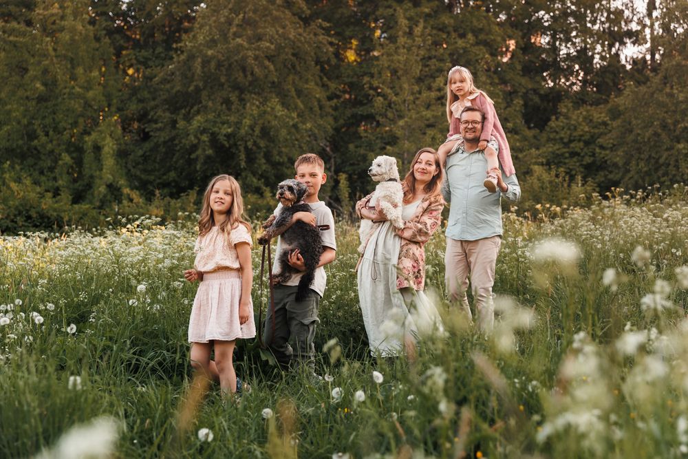 Family with children and dog posing in a blooming meadow with white flowers and lush green trees in background.
