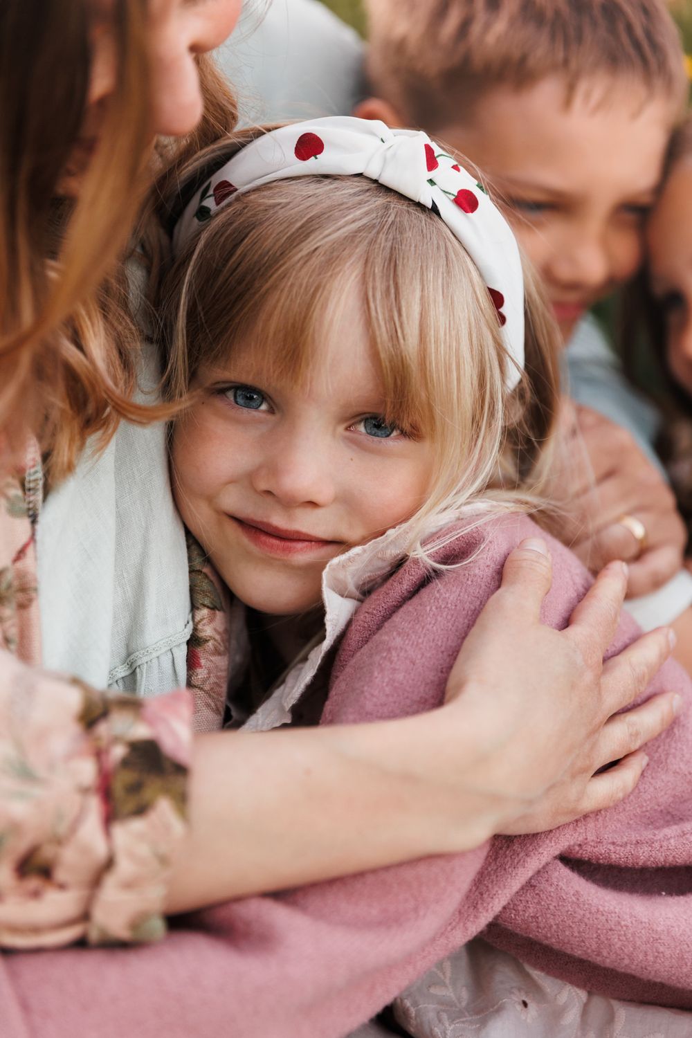 A smiling blonde girl with blue eyes wearing a floral headband is hugged by her family outdoors.