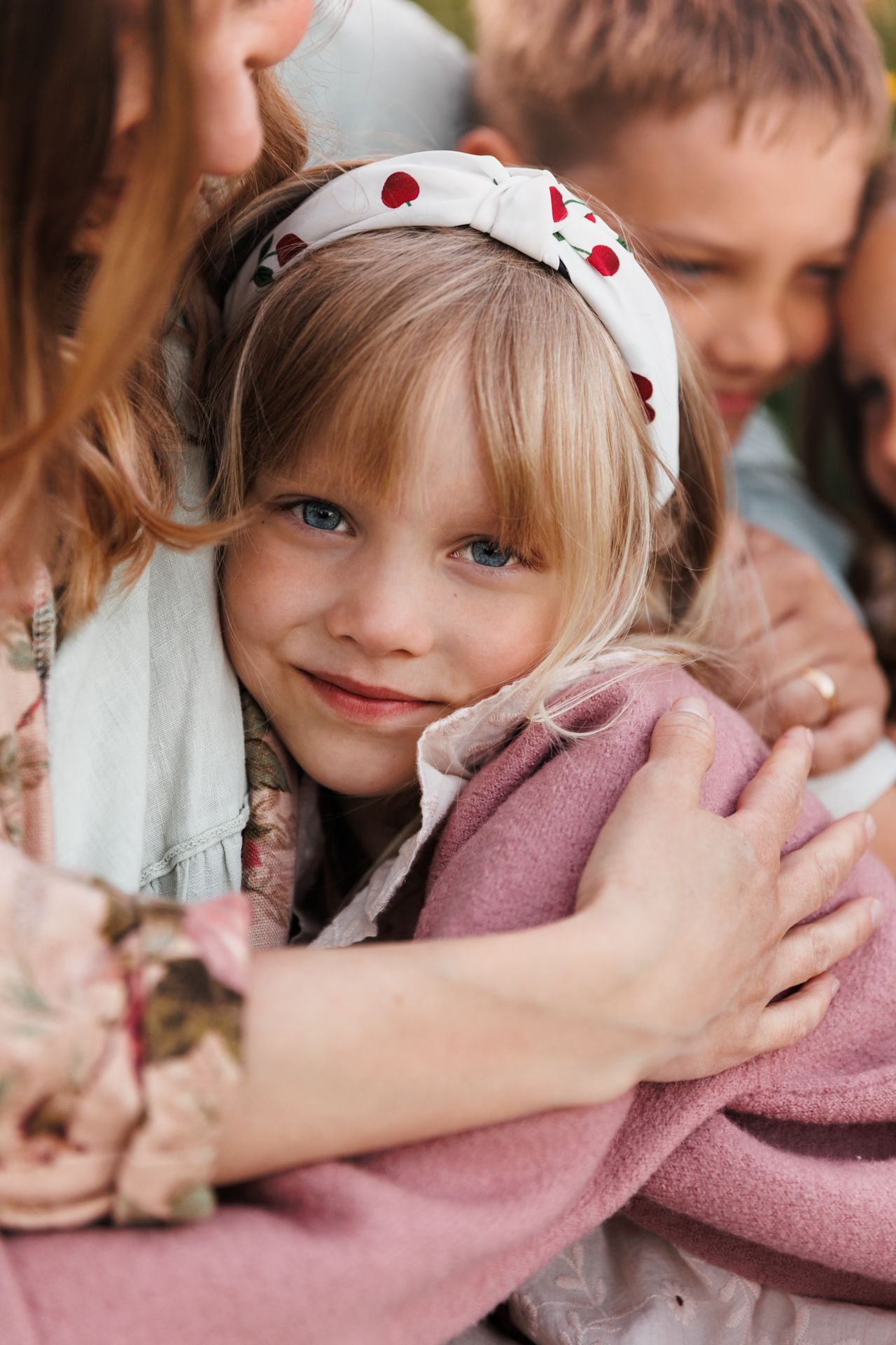 A smiling blonde girl with blue eyes wearing a floral headband is hugged by her family outdoors.
