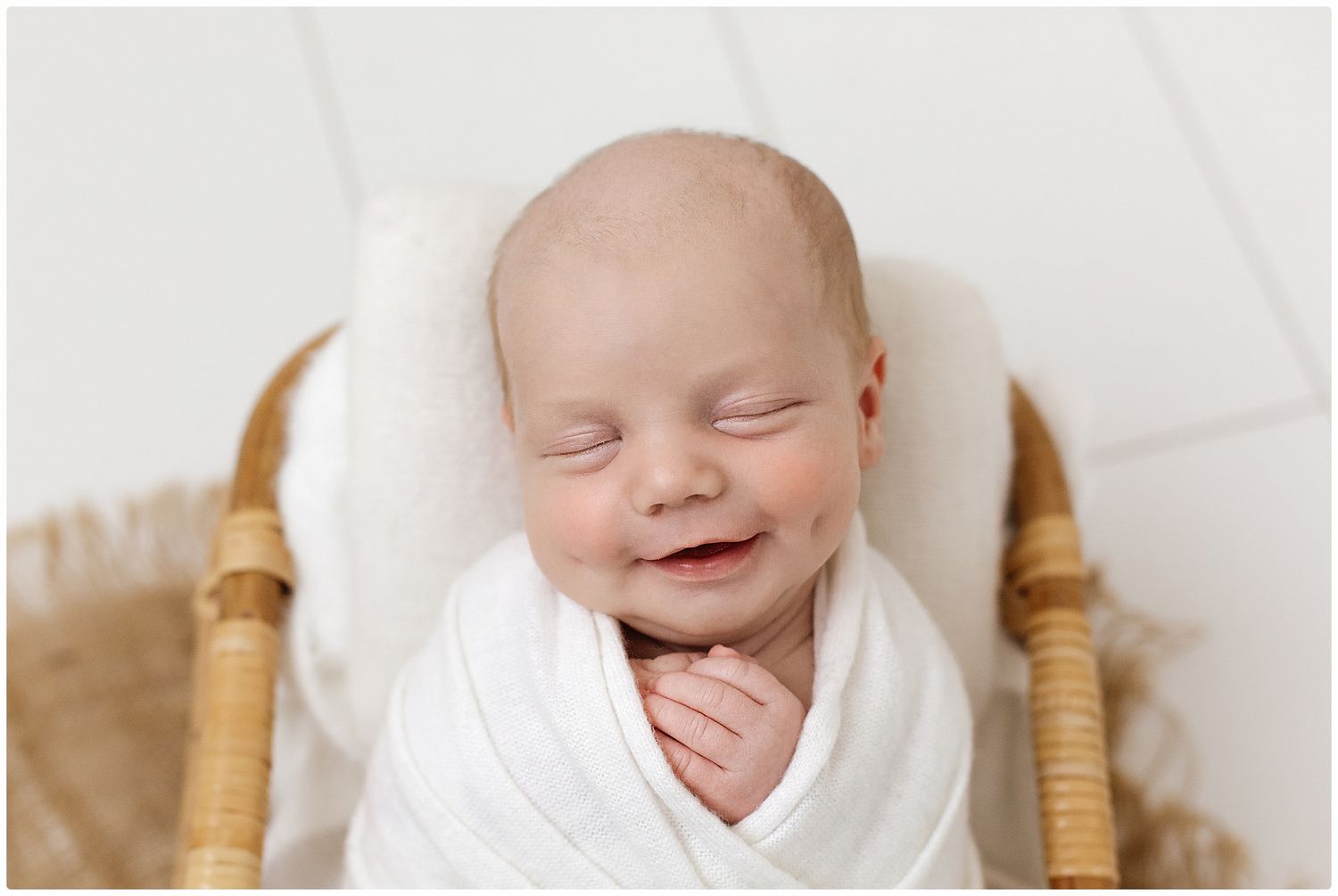Adorable smiling newborn baby wrapped in white swaddle sitting in a wicker basket on white background.