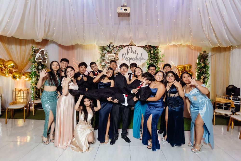 Wedding party guests pose together in a festive reception tent with white draping and decorative lighting.