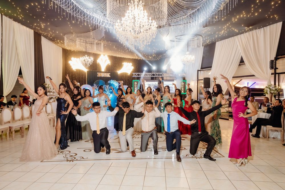 Wedding guests dance together in a large reception hall under elegant crystal chandeliers.