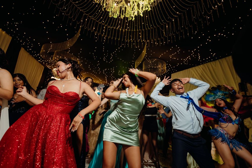 Large group of party guests dancing in formation during a celebration under decorative lighting.