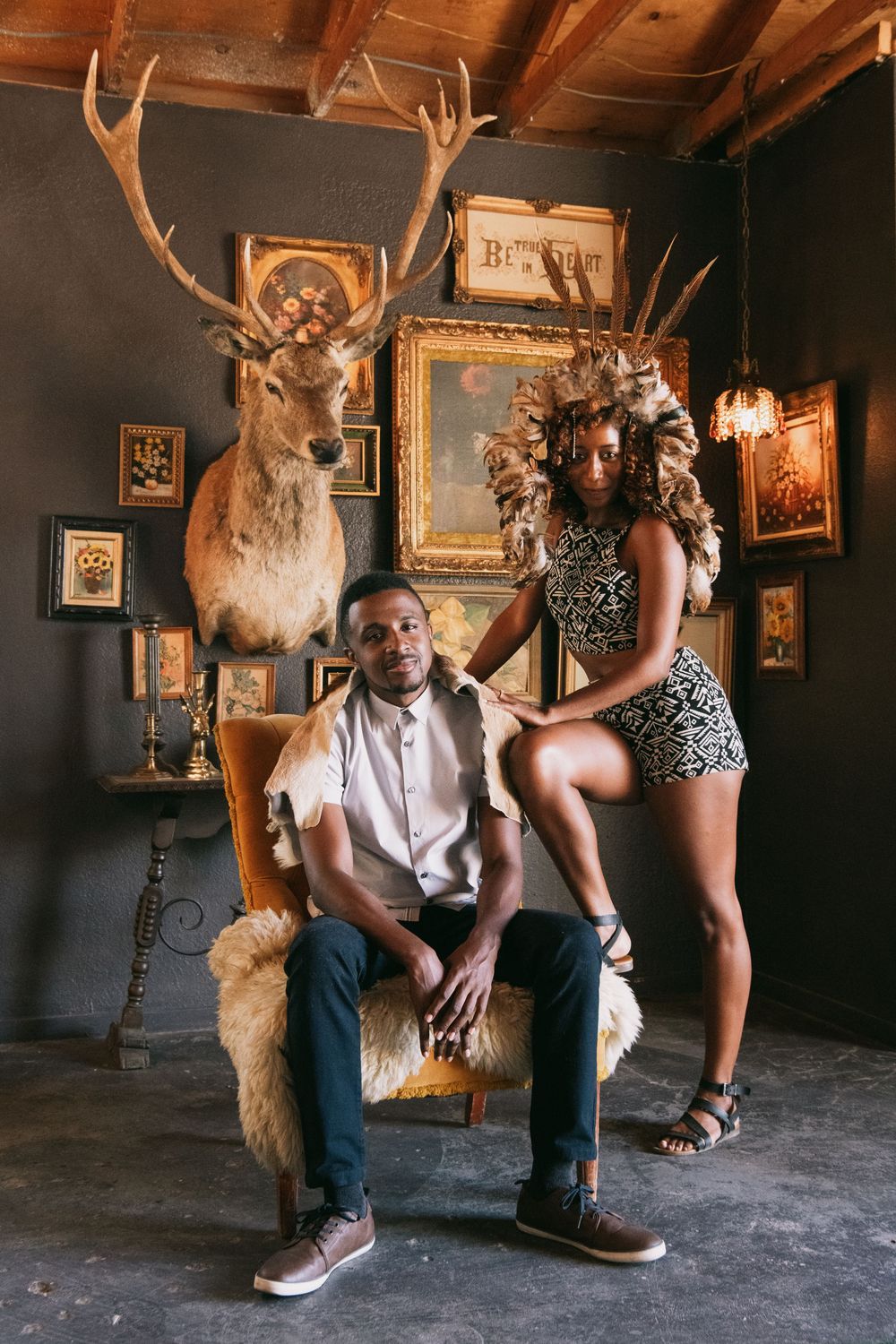 A man and woman pose in a vintage room with taxidermy deer, ornate frames, and fur-covered chair.