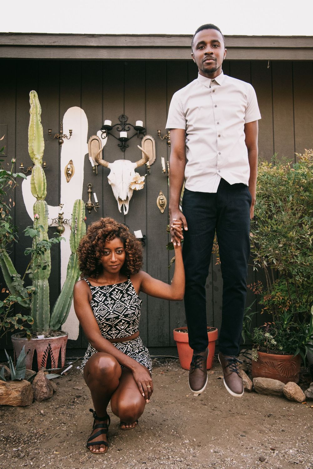 A young Black couple poses outdoors near cacti and a bull skull, woman crouching while man stands holding hands.