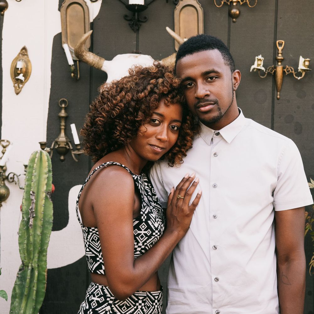 A stylish Black couple poses together in front of a decorative vintage wall with antlers and brass accents.