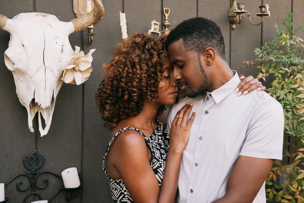 A loving Black couple shares an intimate moment outdoors, embracing near a rustic wall with a decorative animal skull.