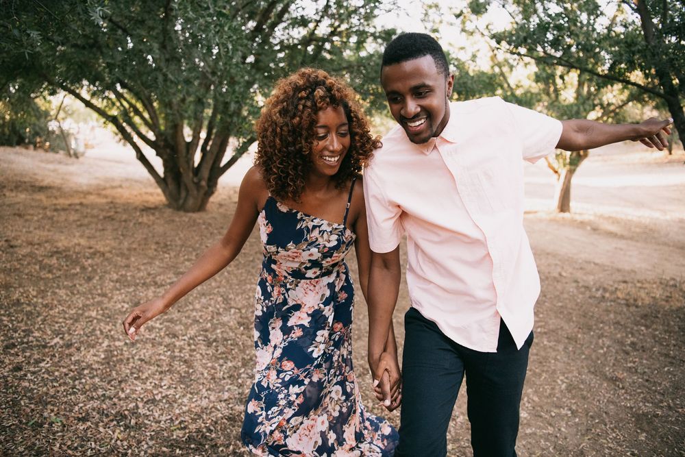 A joyful Black couple holds hands and dances together outdoors among trees, woman in floral dress, man in pink shirt.
