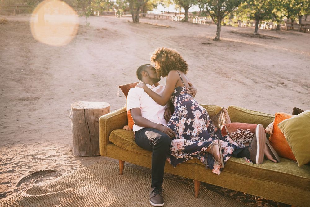 A couple embraces warmly on a vintage sofa outdoors, surrounded by sandy ground and trees in soft sunlight.