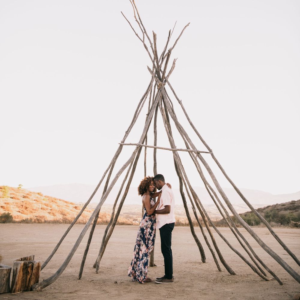 Couple embracing under a rustic wooden teepee structure in a desert landscape at dusk.