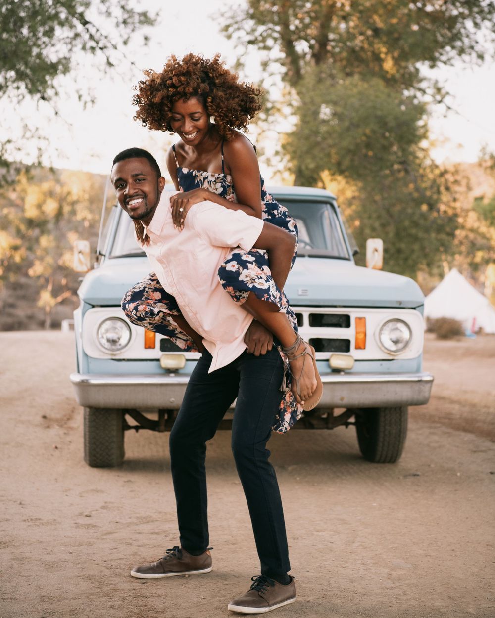 A joyful man gives a woman a piggyback ride in front of a vintage blue truck on a sunny dirt road.