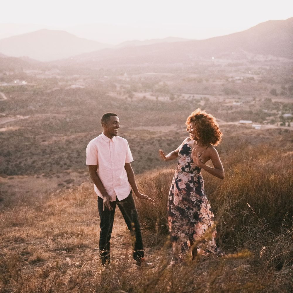 A couple shares a joyful moment on a sunlit hillside with scenic mountain valley views in the background.