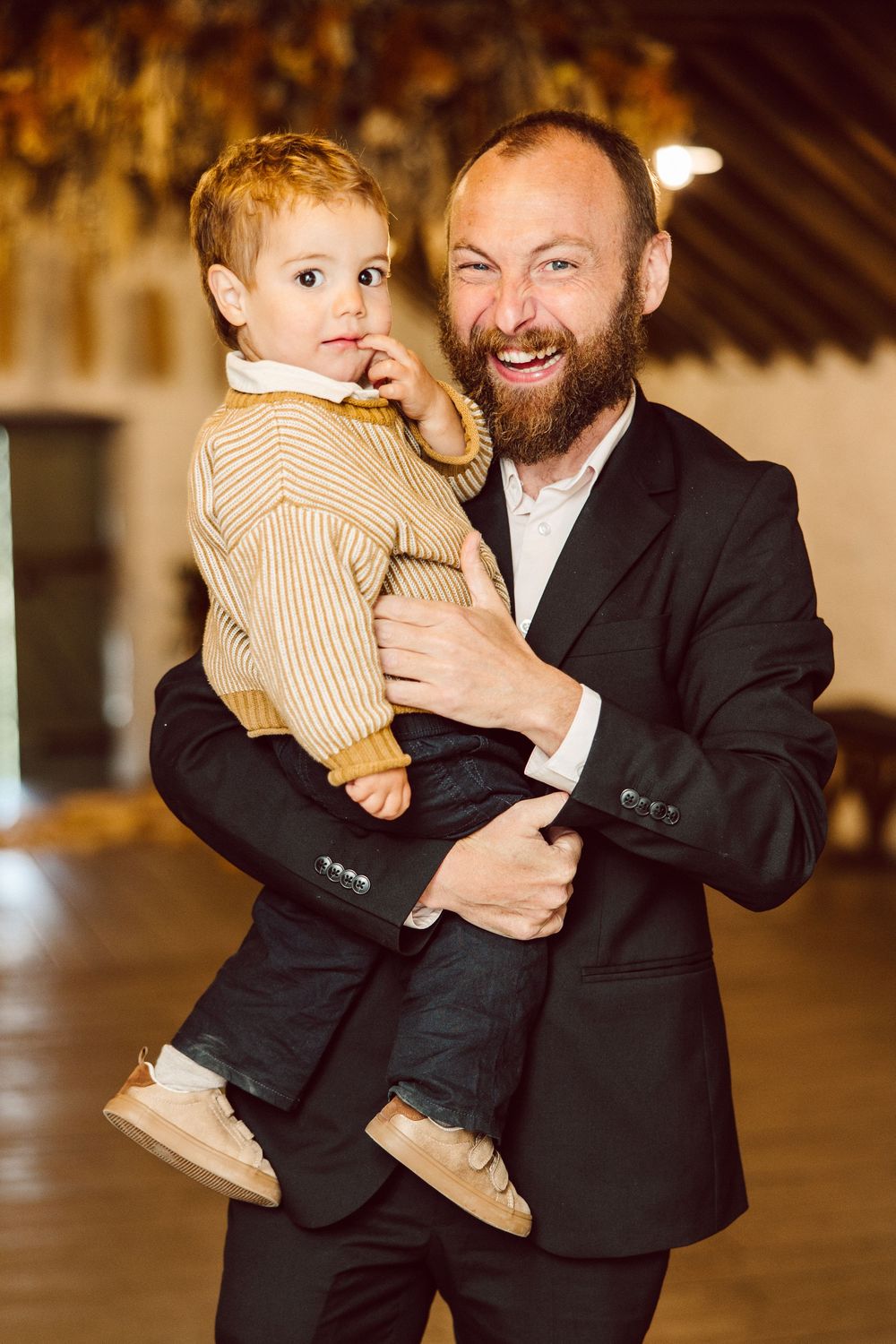 A man in a black suit holds a small child wearing a striped outfit while dancing at an indoor venue.
