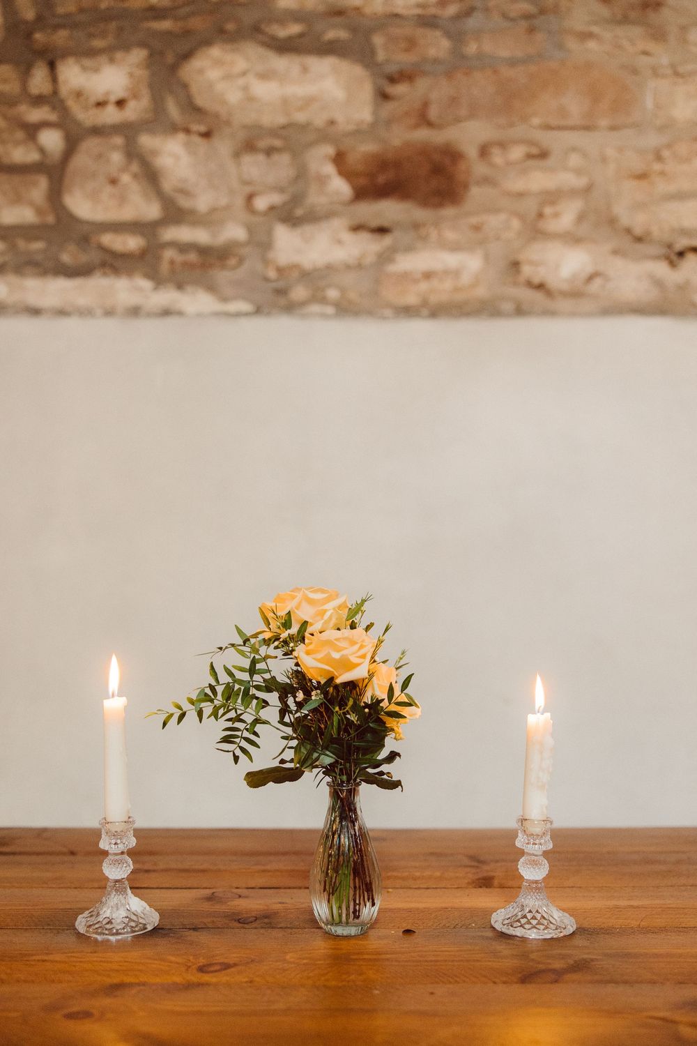 A rustic wooden table with white candles and yellow roses in a glass vase against a stone wall.