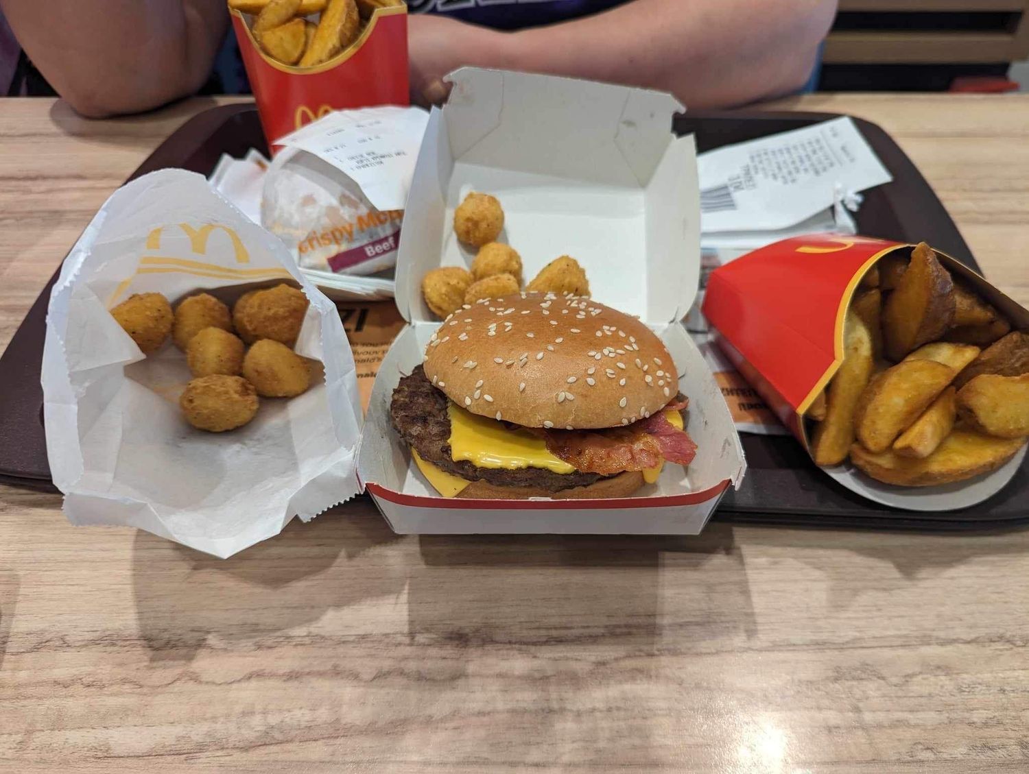 A McDonald's meal spread on a wooden table featuring a cheeseburger, chicken nuggets, and potato wedges with packaging.