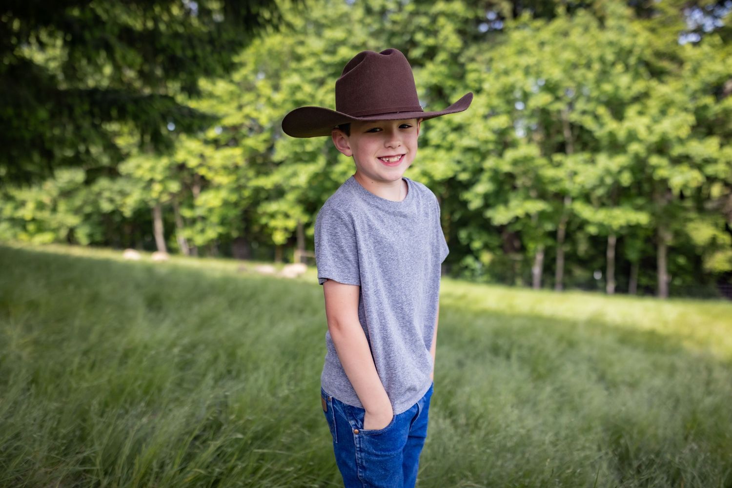 Smiling boy wearing a brown cowboy hat and gray t-shirt stands in a lush green field.