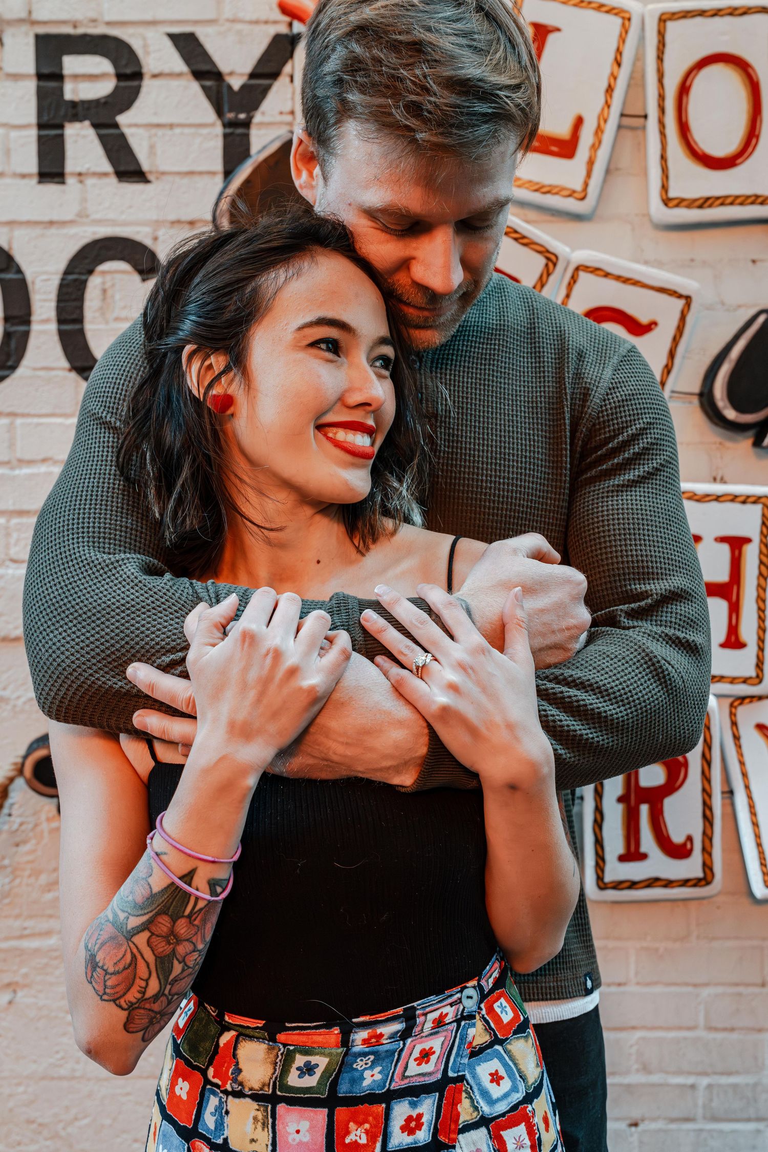 Smiling tattooed woman being embraced by man in front of decorative sign wall.