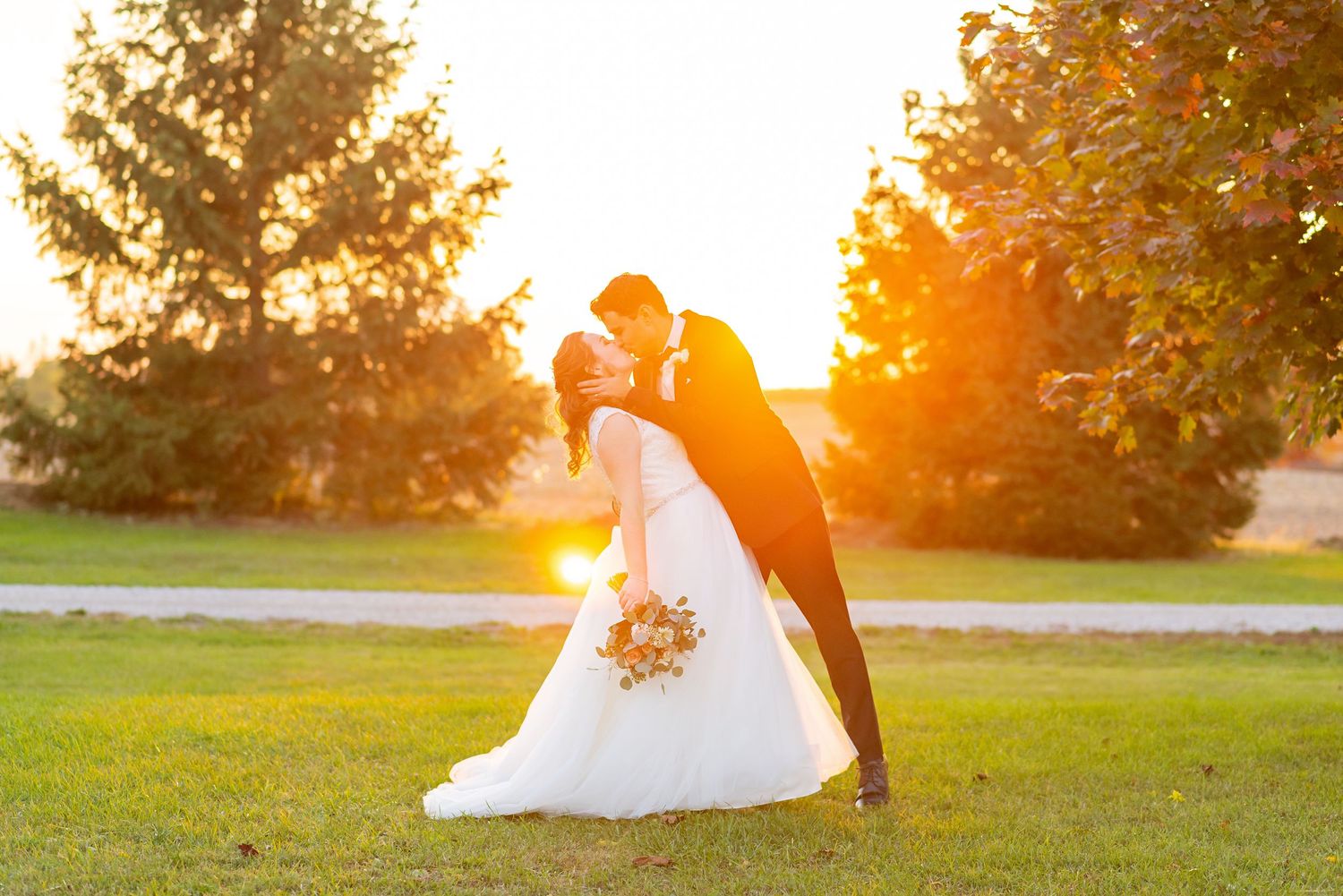 A couple embraces during golden hour in a grassy field with sunlight streaming through trees in the background.