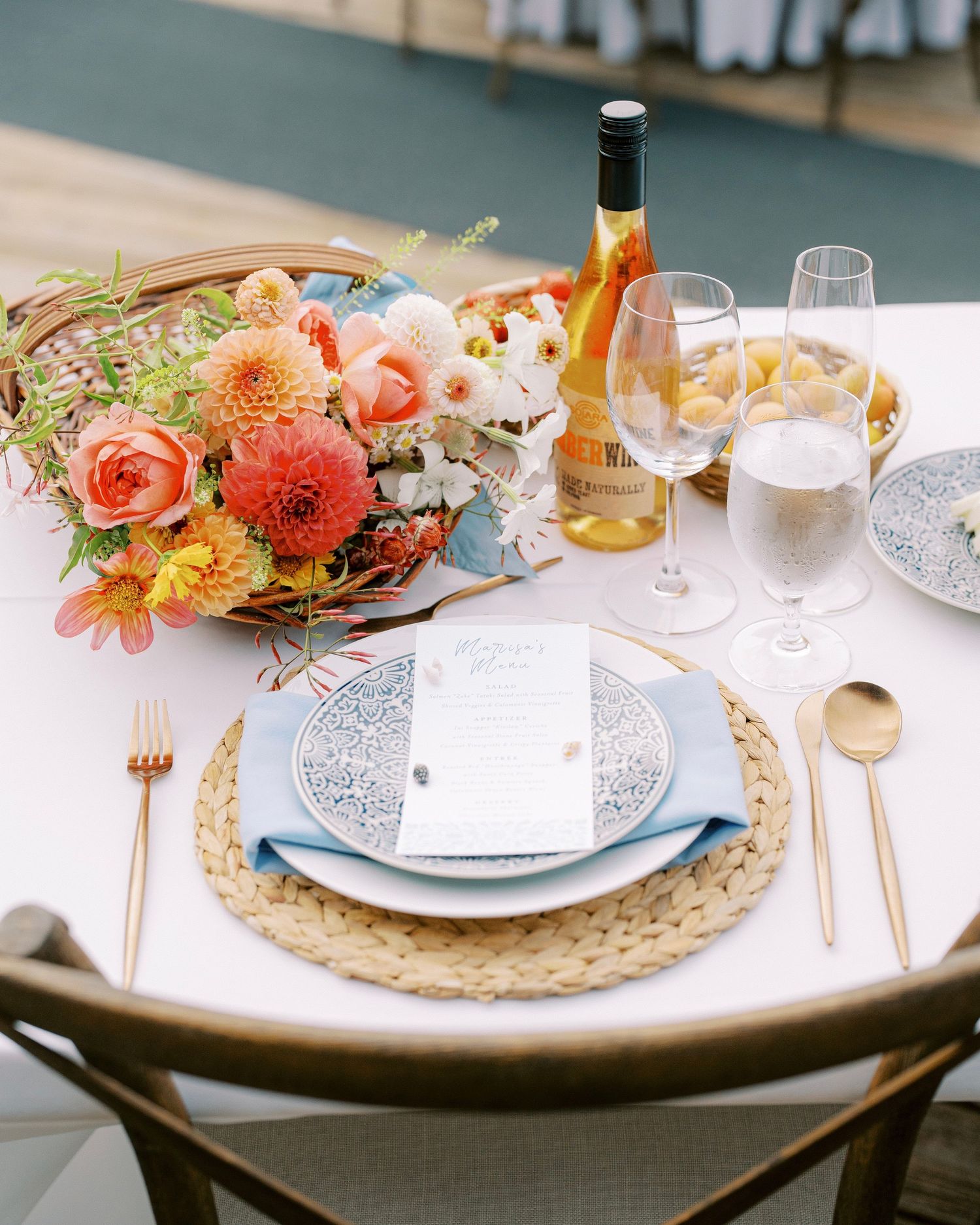 Romantic table setting with coral flowers, gold cutlery, and woven placemats for an outdoor wedding reception.