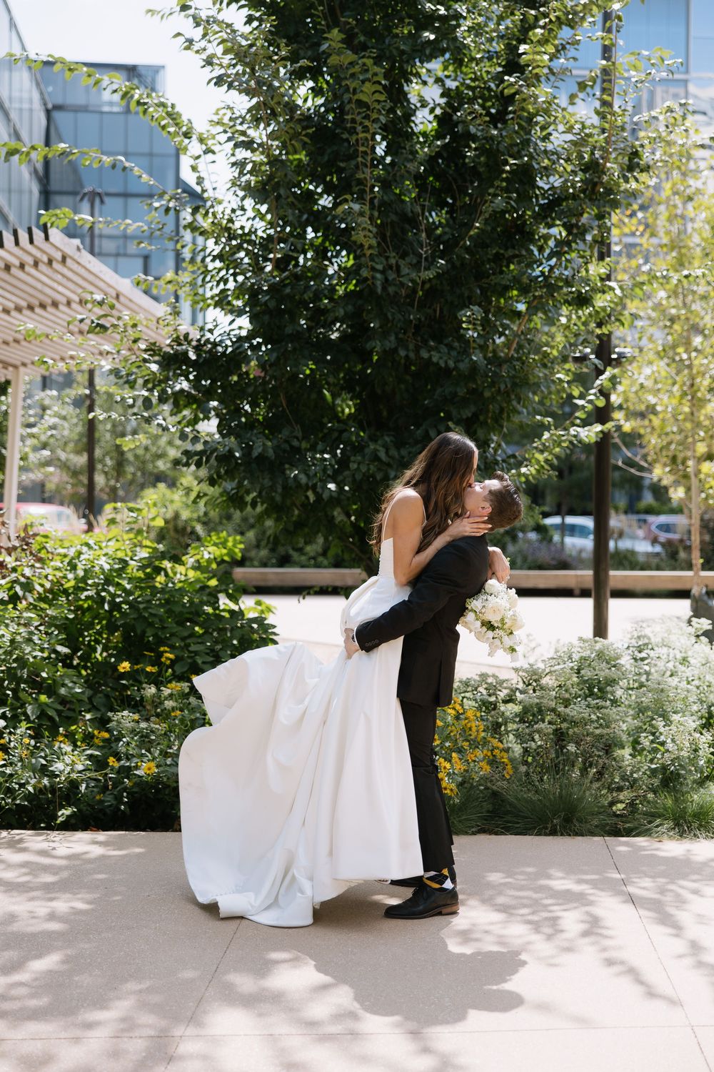 A couple in wedding attire dips dramatically against a backdrop of trees and greenery.