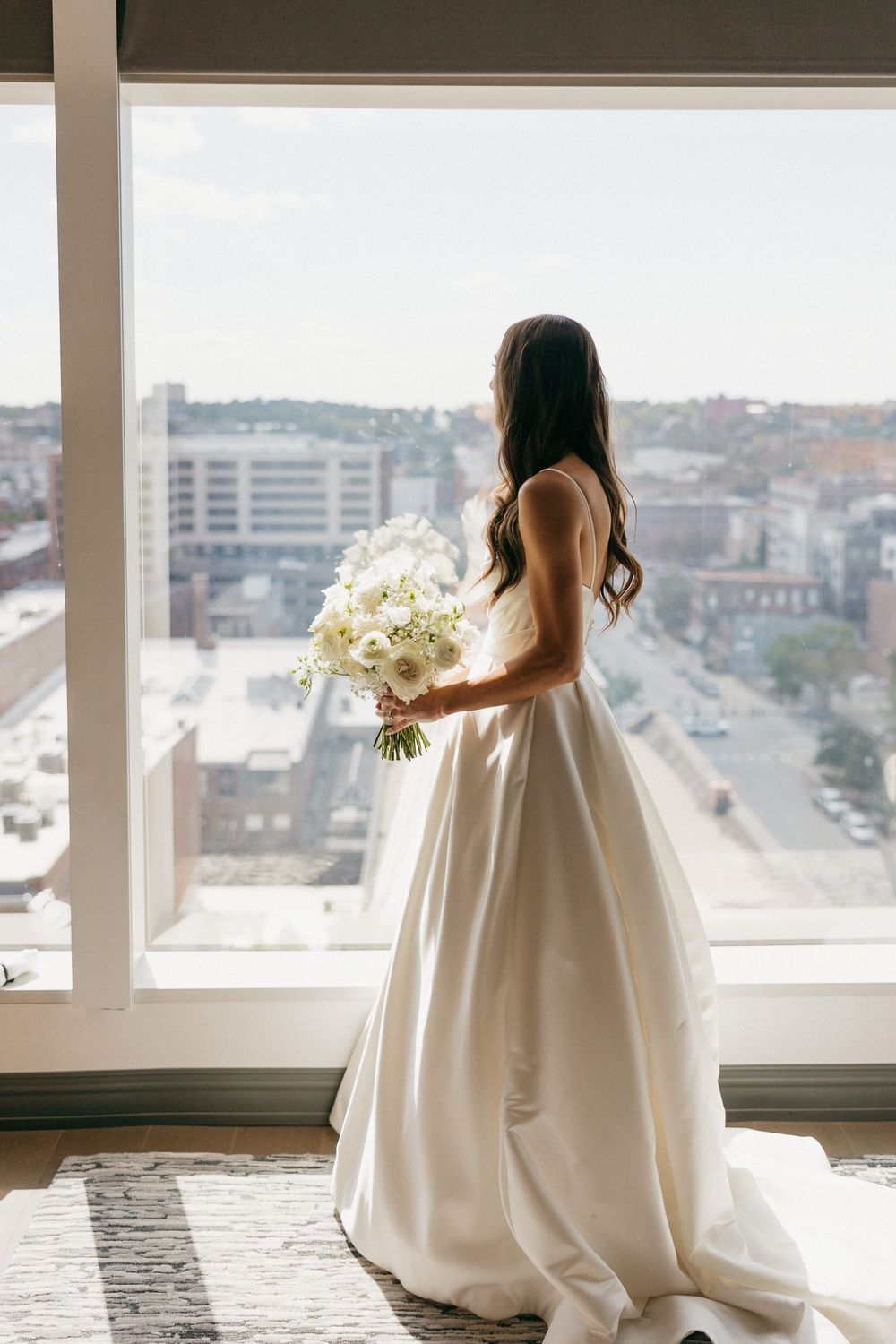 Elegant white wedding dress silhouetted against large windows with urban backdrop.