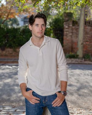 Person in white collared shirt and jeans sitting on rustic stone wall in dappled sunlight.