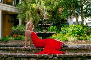 A women in a flowing red dress poses gracefully across a brick wall in a lush garden setting in Downtown Charleston, SC.