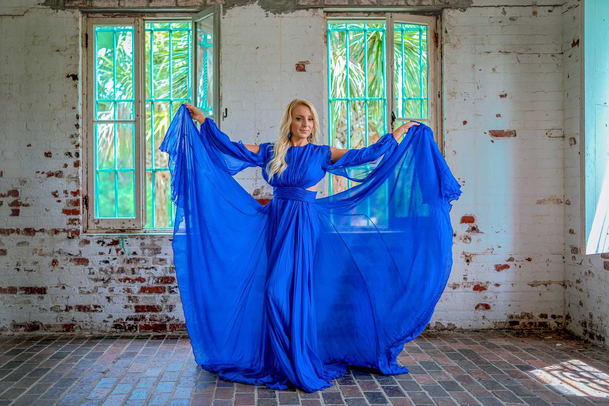 Model portrait in Charleston, SC royal blue dress billows dramatically in an abandoned industrial space with sunlit windows.