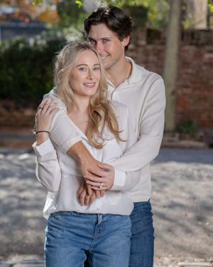 A romantic couples portrait in matching white sweaters and jeans embrace outdoors against a brick wall backdrop at Downtown Charleston, SC.