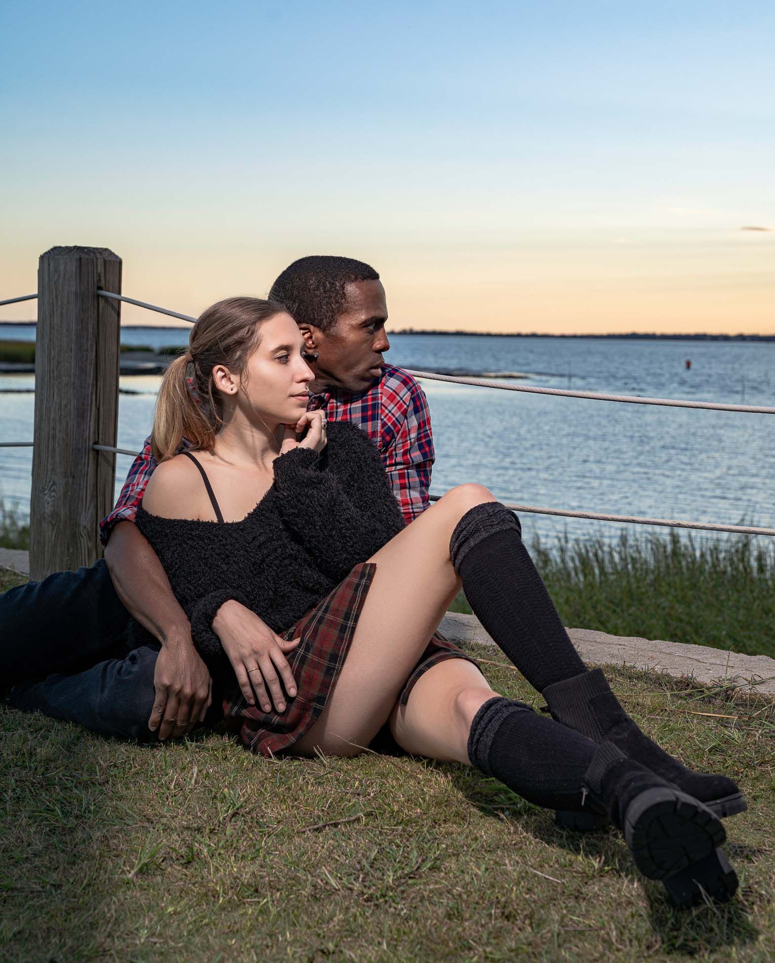Couple portrait together on grassy shore at sunset with water and bridge view in background at Pitt Street Bridge, Mount Pleasant, SC.
