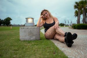 A model in fishnet stockings and black boots poses on a brick path near grass and palm trees.