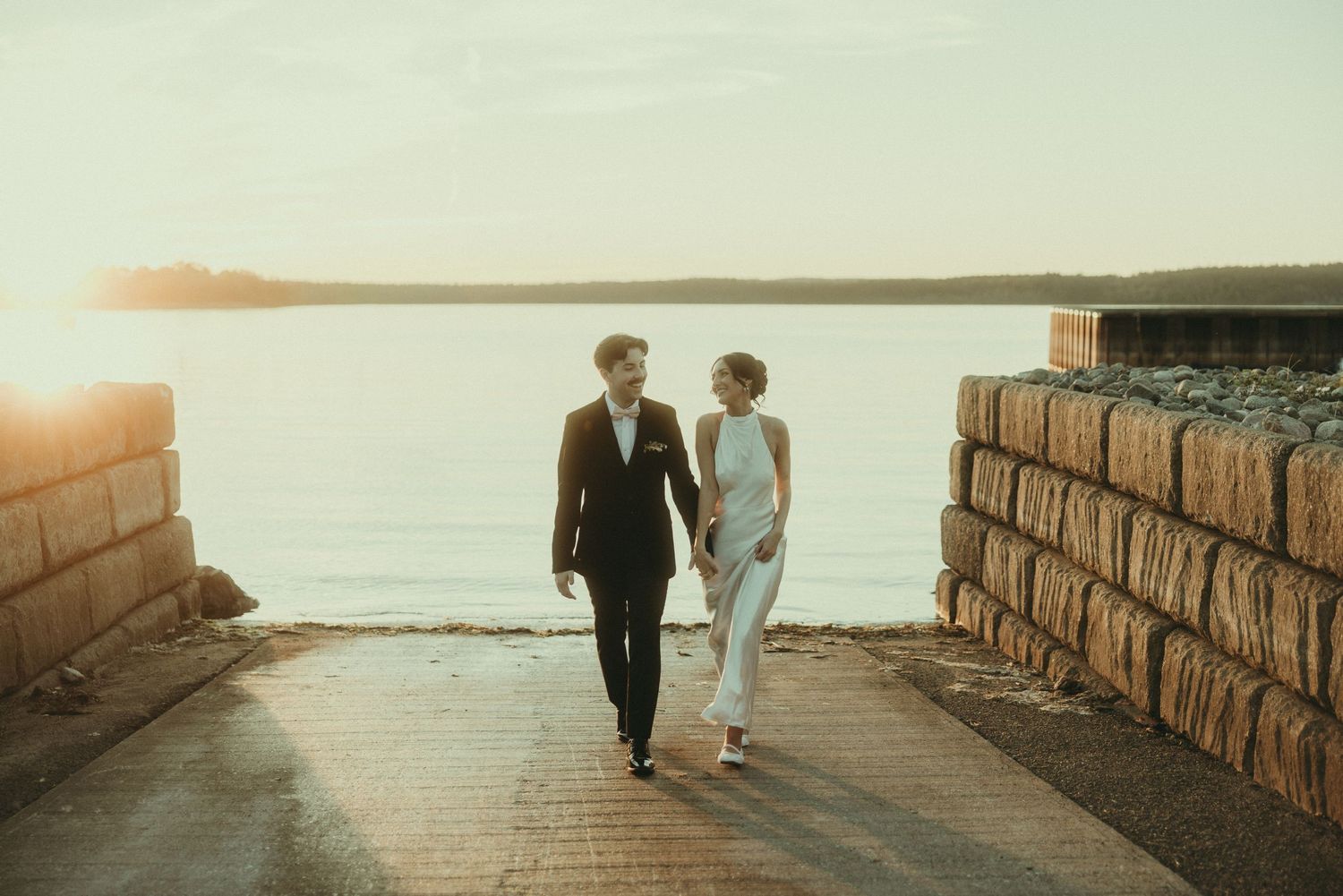 Silhouettes of a couple walking along a stone wall by the ocean at sunset.