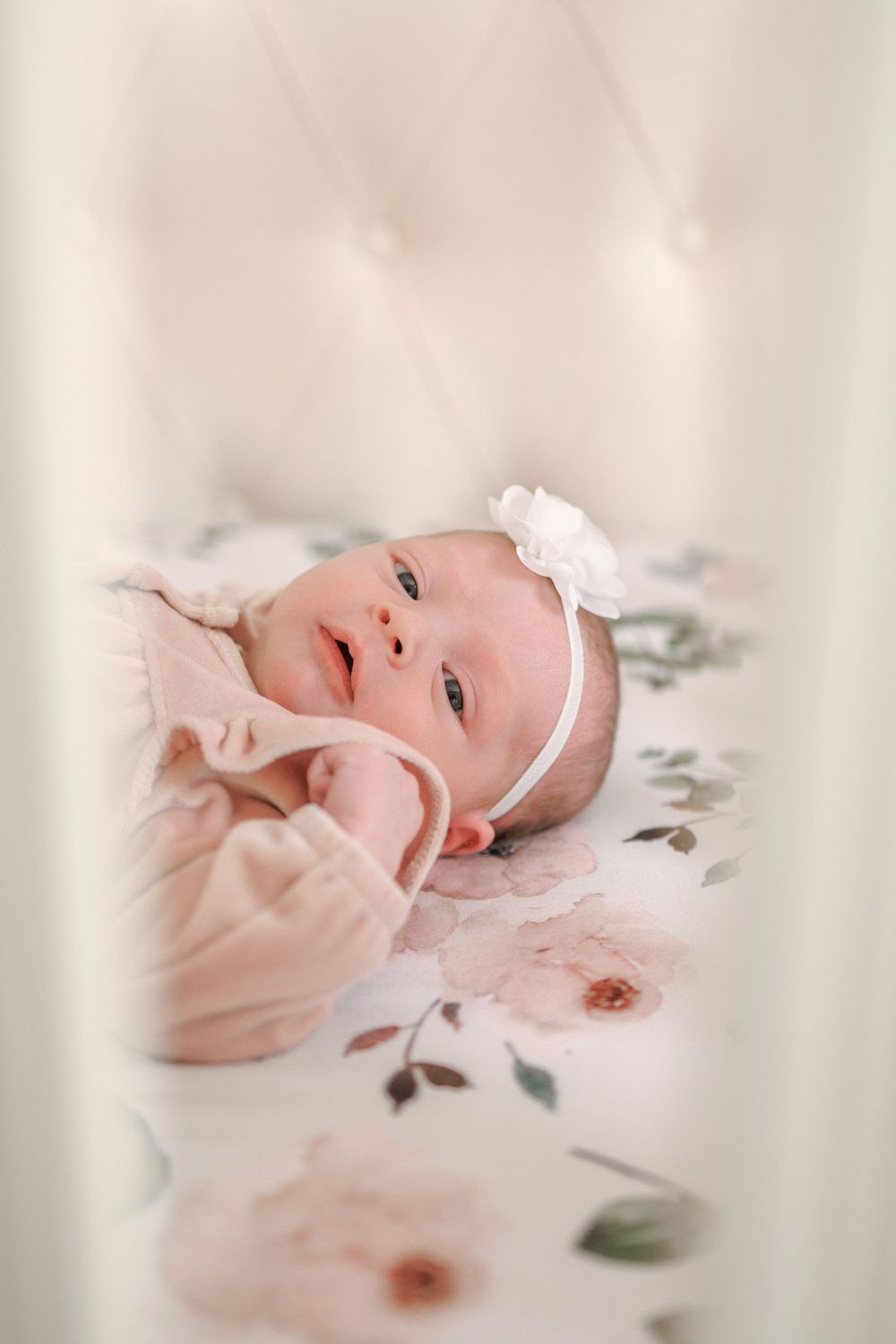 A newborn baby wearing a white headband lies on floral bedding in soft pink tones.
