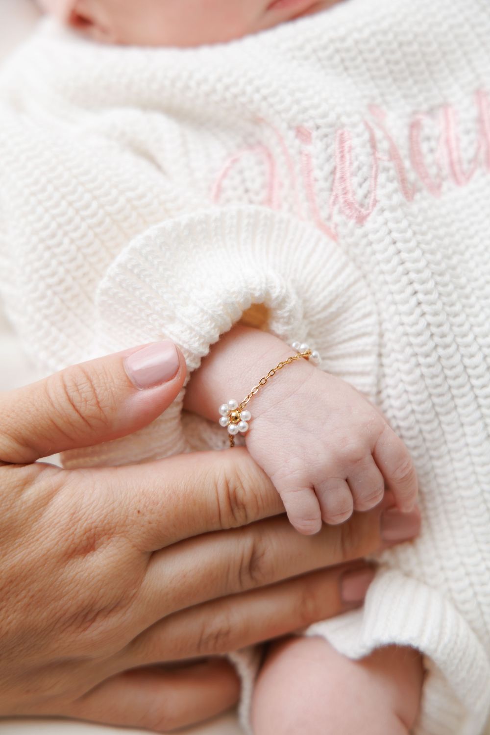 A tiny baby's hand wearing a delicate gold daisy bracelet against a white knitted sweater background.