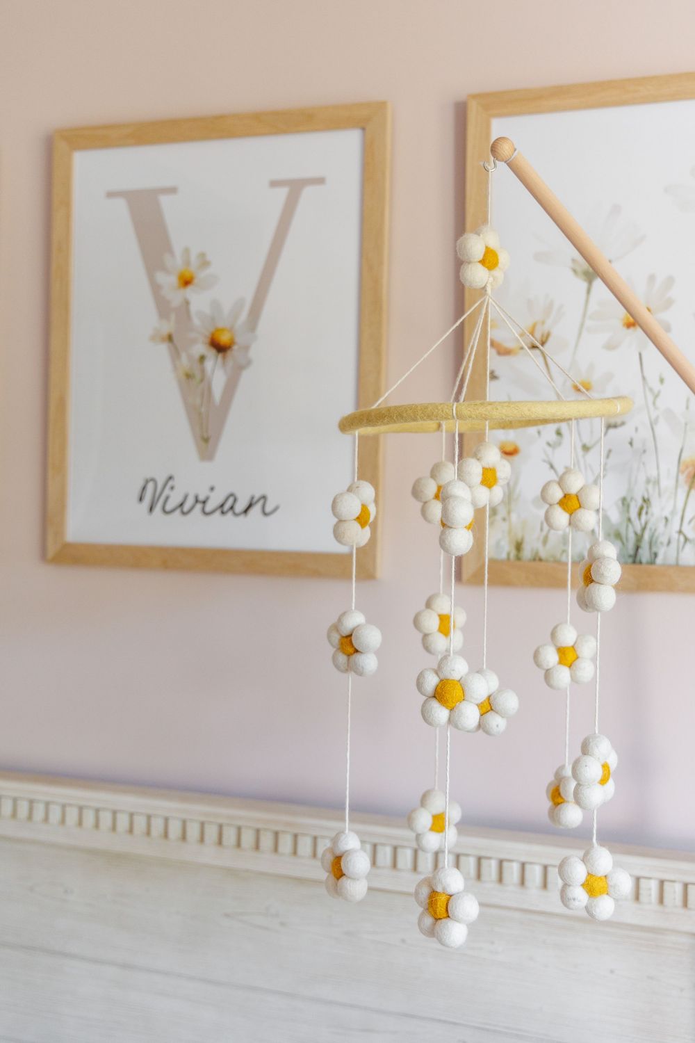 A delicate white and yellow daisy mobile hangs from the ceiling against a light neutral wall with framed artwork.