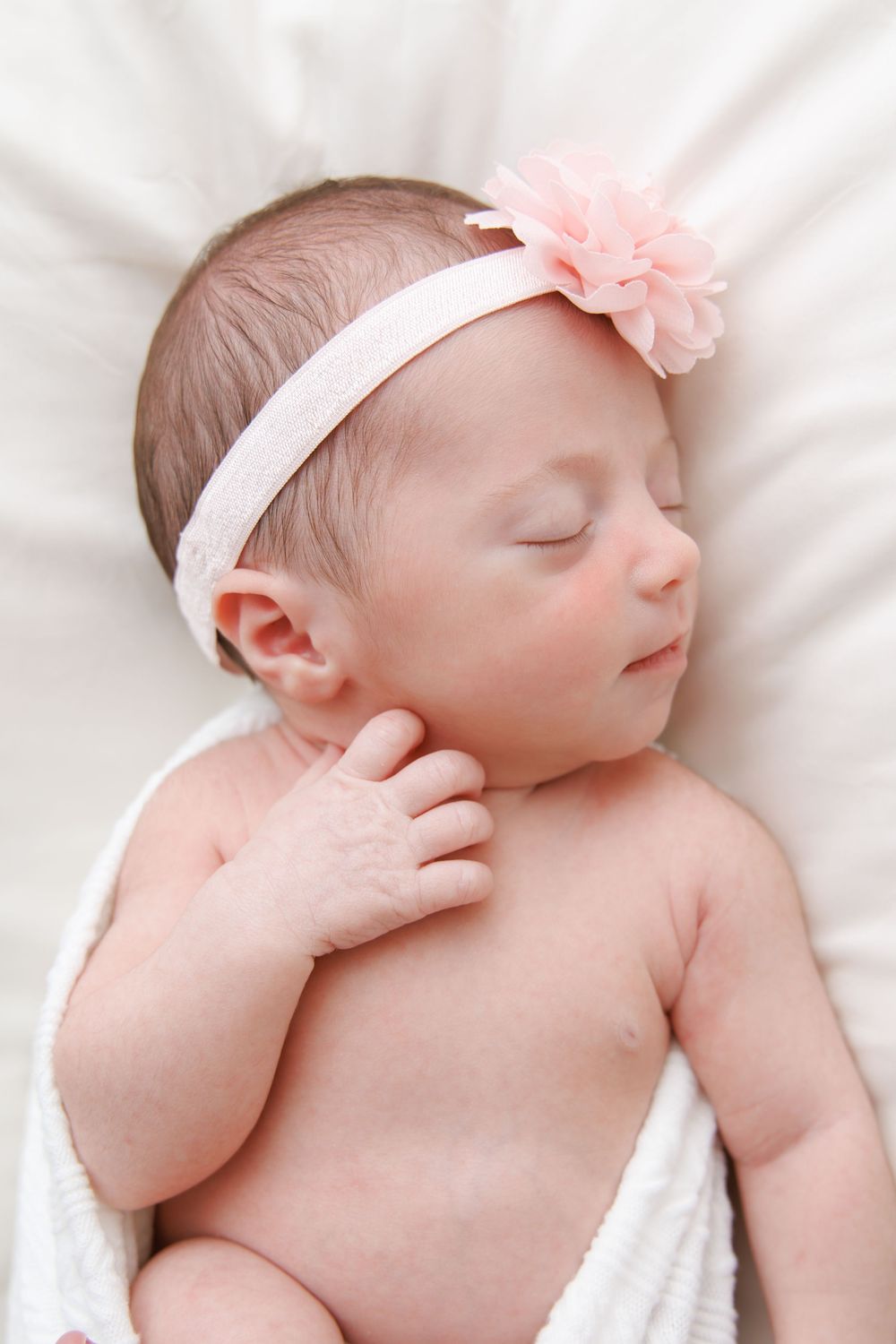 A sleeping newborn baby wearing a pink bow headband lies peacefully on white bedding.