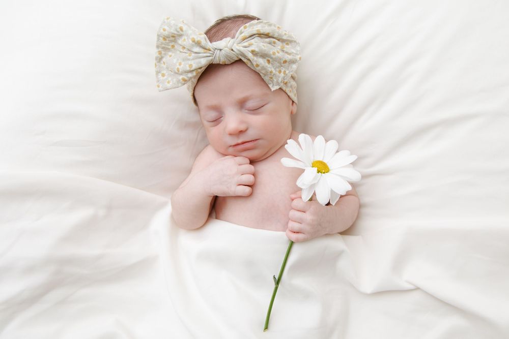 Sleeping newborn wearing lace bonnet lies on white blanket next to single daisy flower.