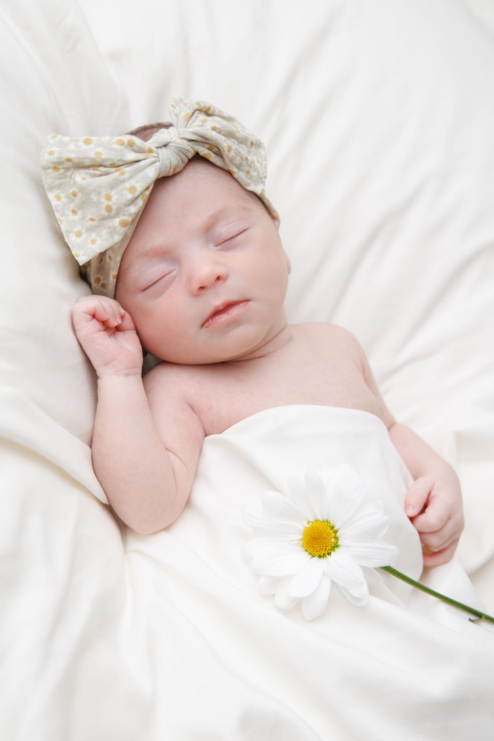 Sleeping newborn in polka dot bow headband with white daisy flower on white bedding.