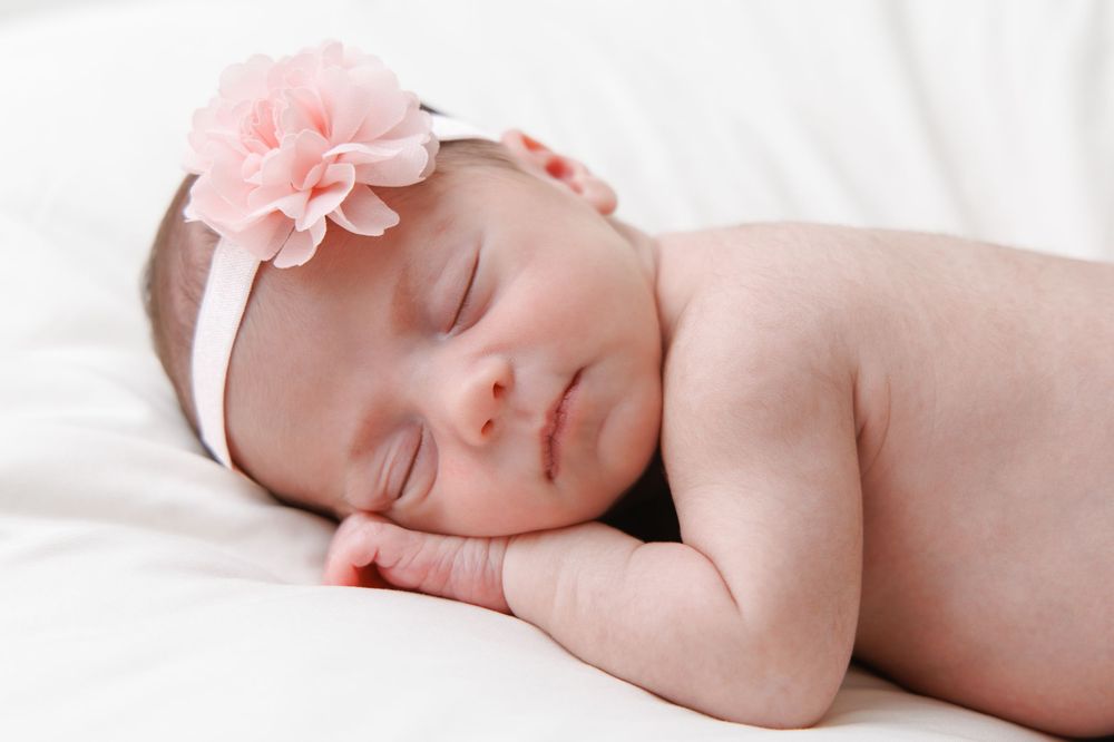 Peaceful sleeping newborn baby wearing pink flower headband on white blanket.