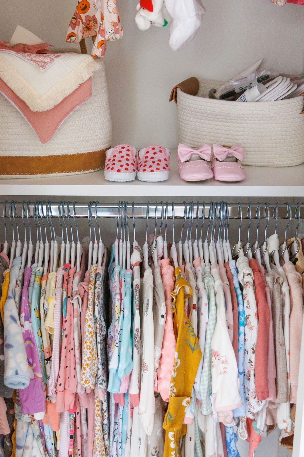 Organized baby nursery closet with pink and floral dresses hanging neatly and tiny shoes displayed on shelf above.