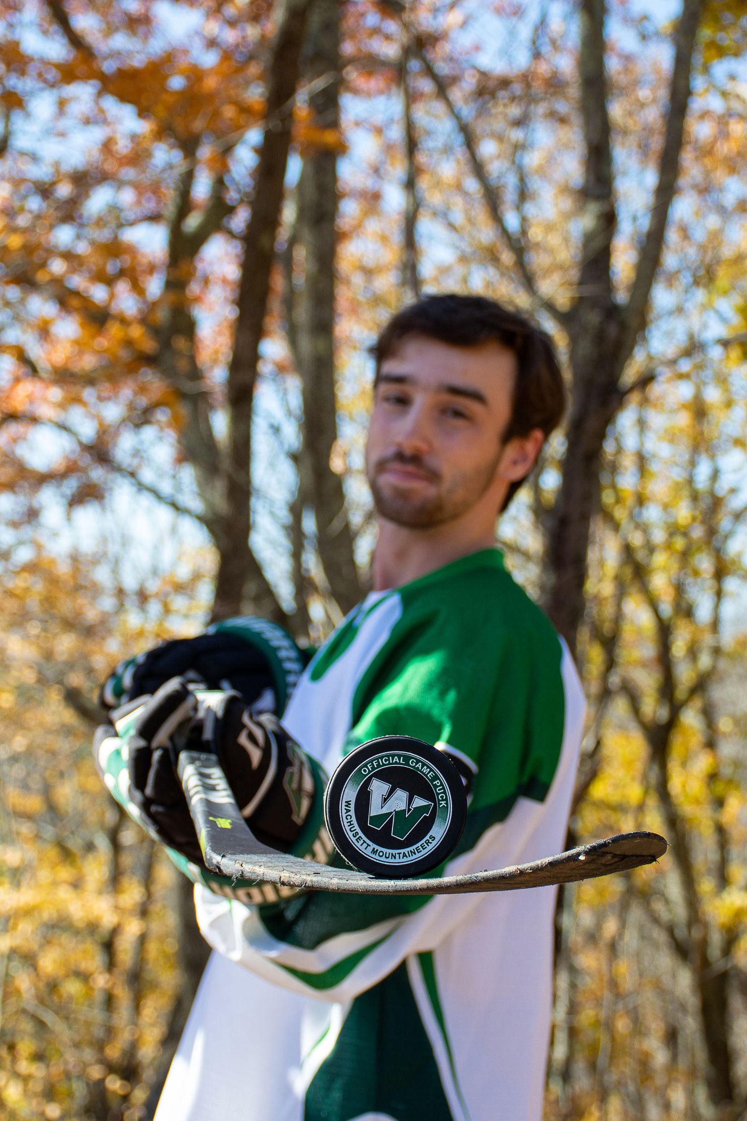 Hockey player in green and white jersey holds stick against autumn forest backdrop during outdoor practice session.