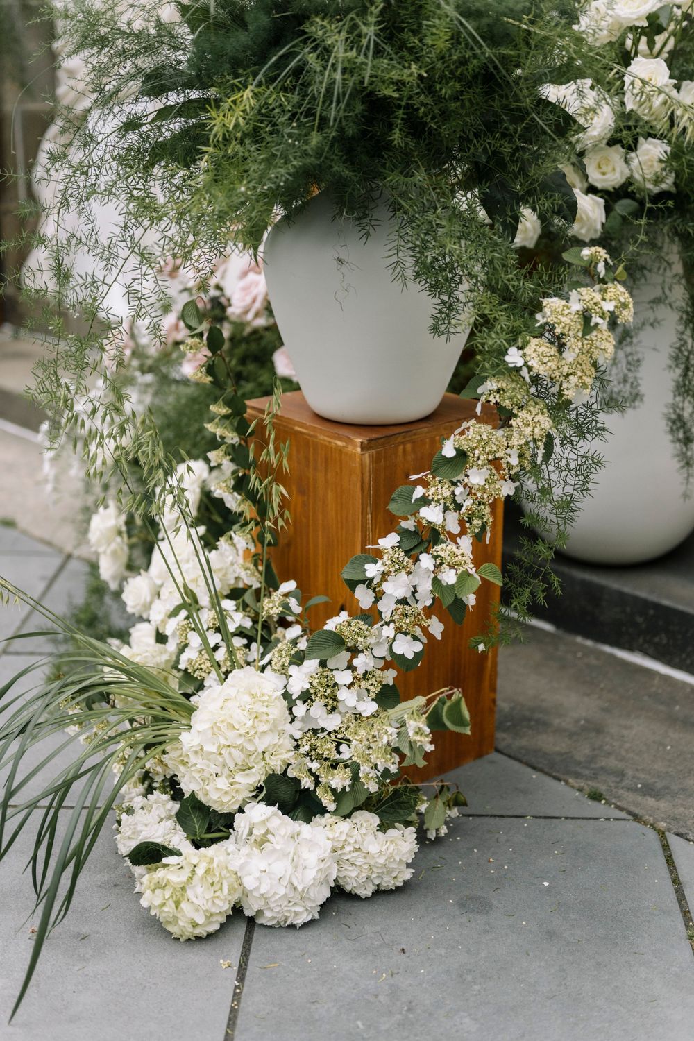Elegant floral arrangement with white hydrangeas and delicate greenery cascading from white planters on wooden pedestal.